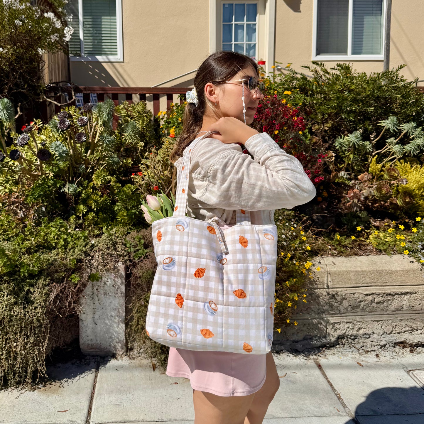Woman holding a patterned tote bag with coffee, croissants and gingham, in front of a house with plants and flowers.