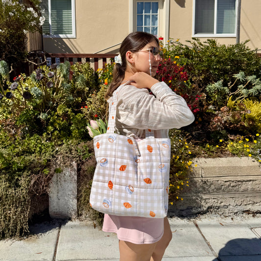Woman holding a patterned tote bag with coffee, croissants and gingham, in front of a house with plants and flowers.